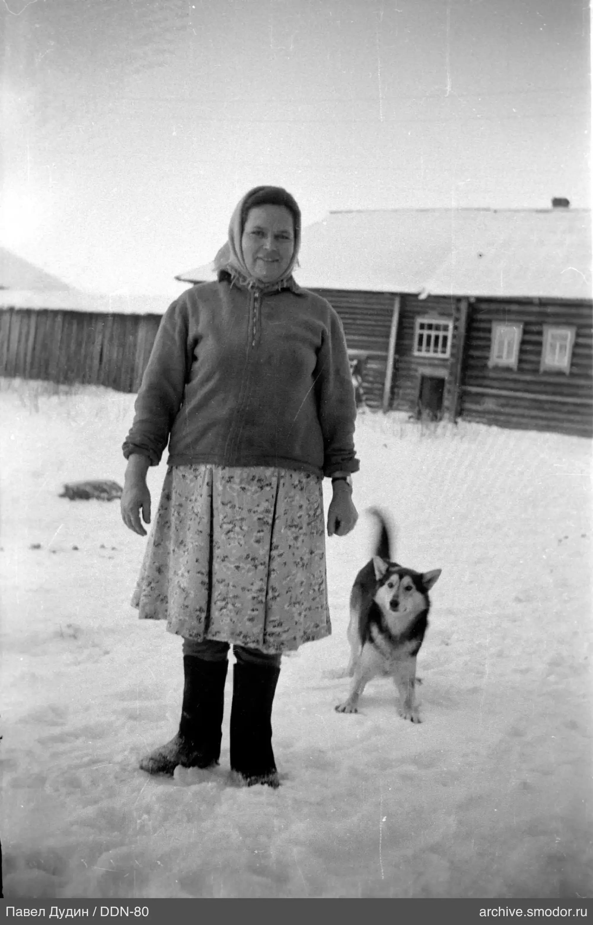 Frau mit Hund im Schnee vor Holzhaus