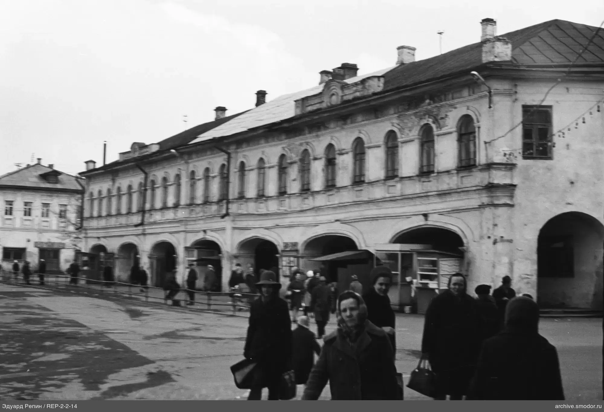 Двухэтажное здание с арками и людьми на площади