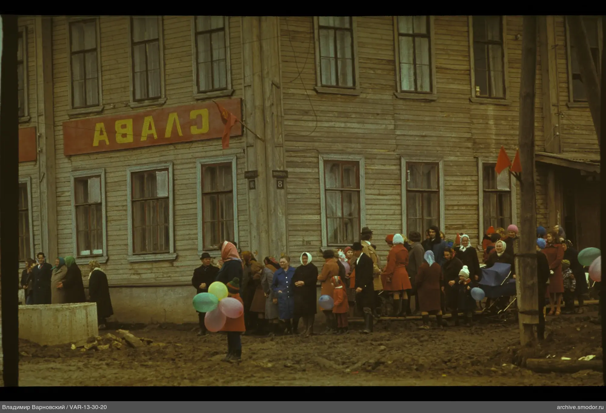 Группа людей у деревянного здания с флагом и вывеской