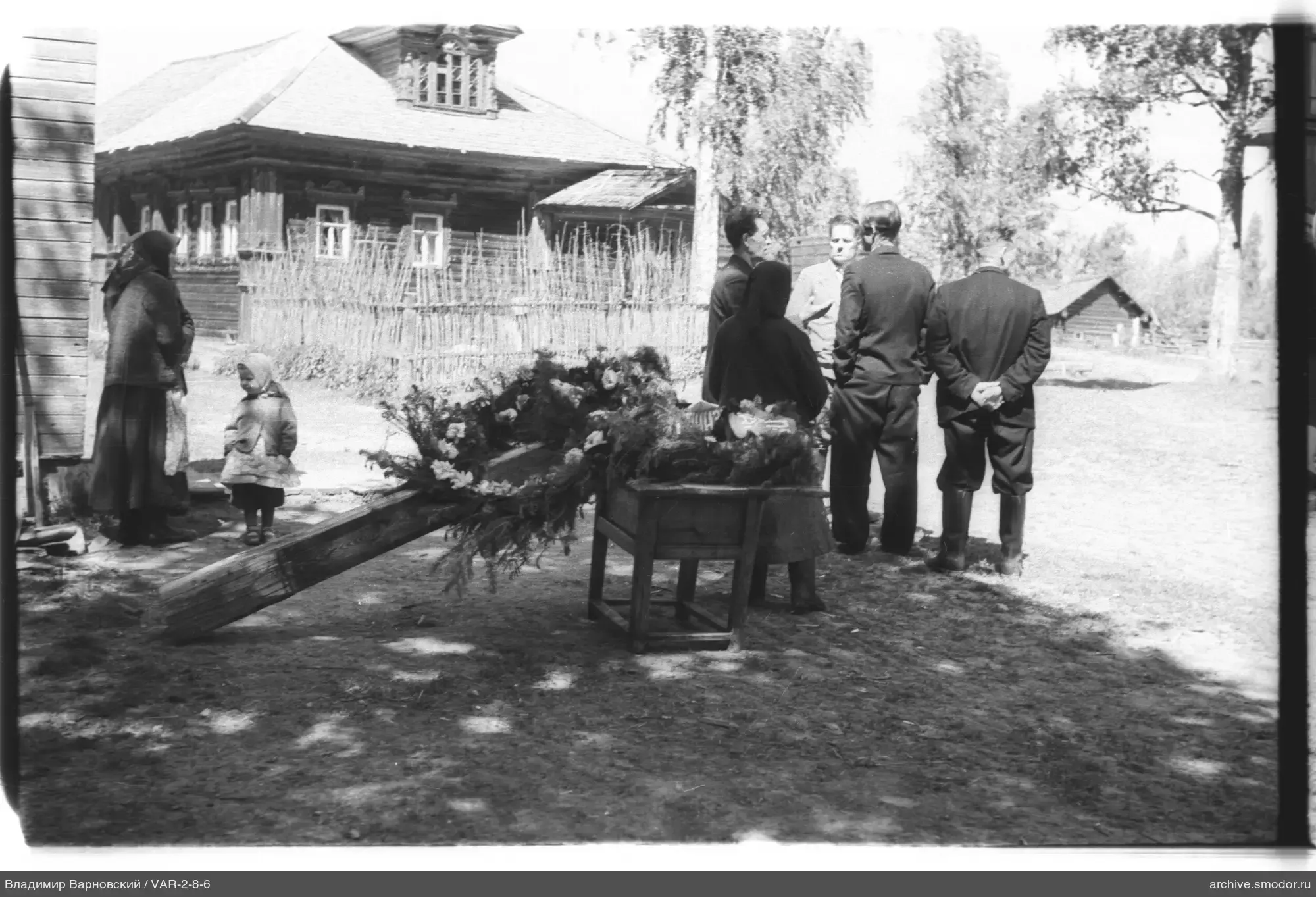 Мужчины и женщины у деревянного дома с носилками