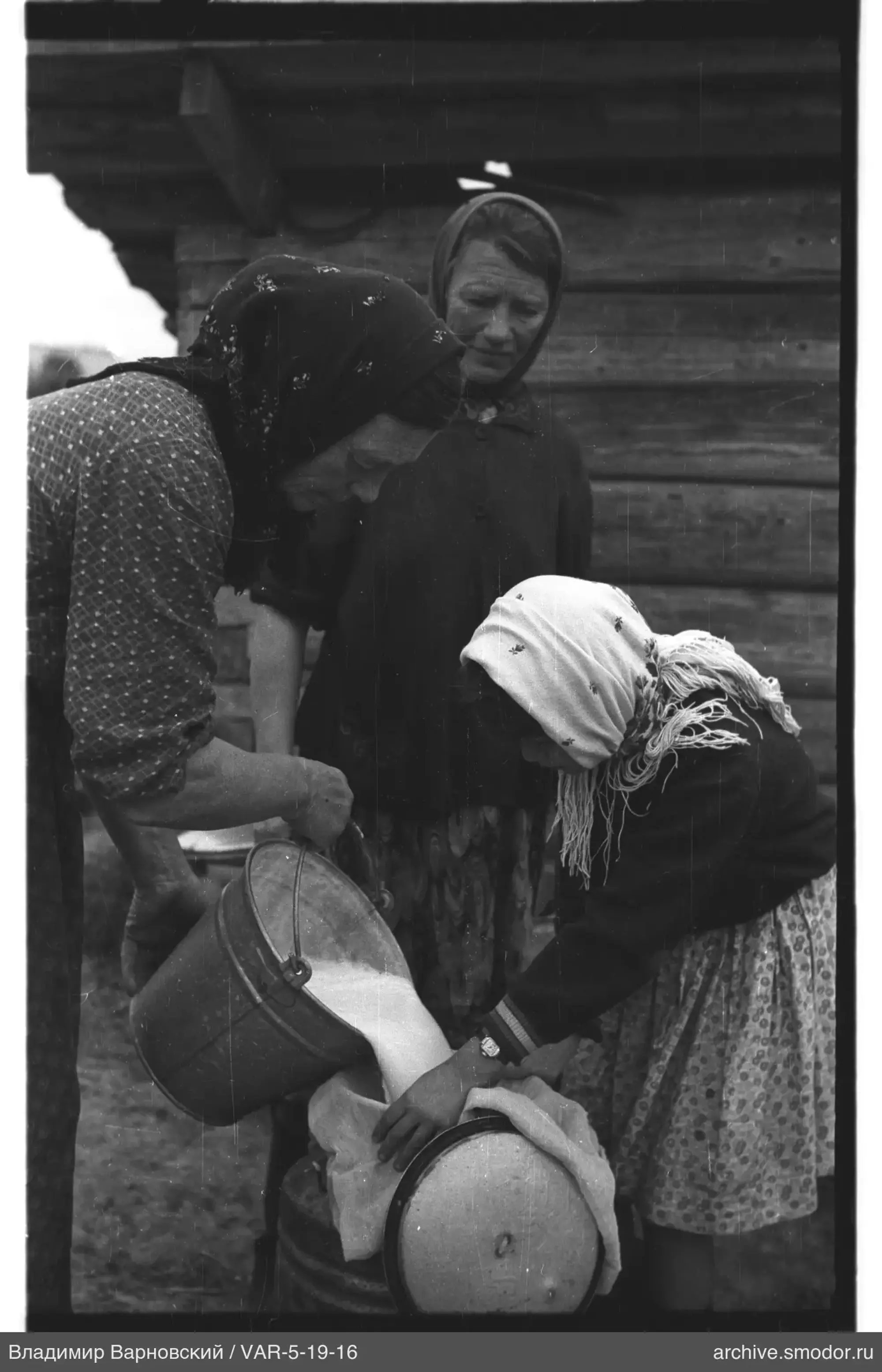 Frauen beim Milch gießen vor Holzhaus