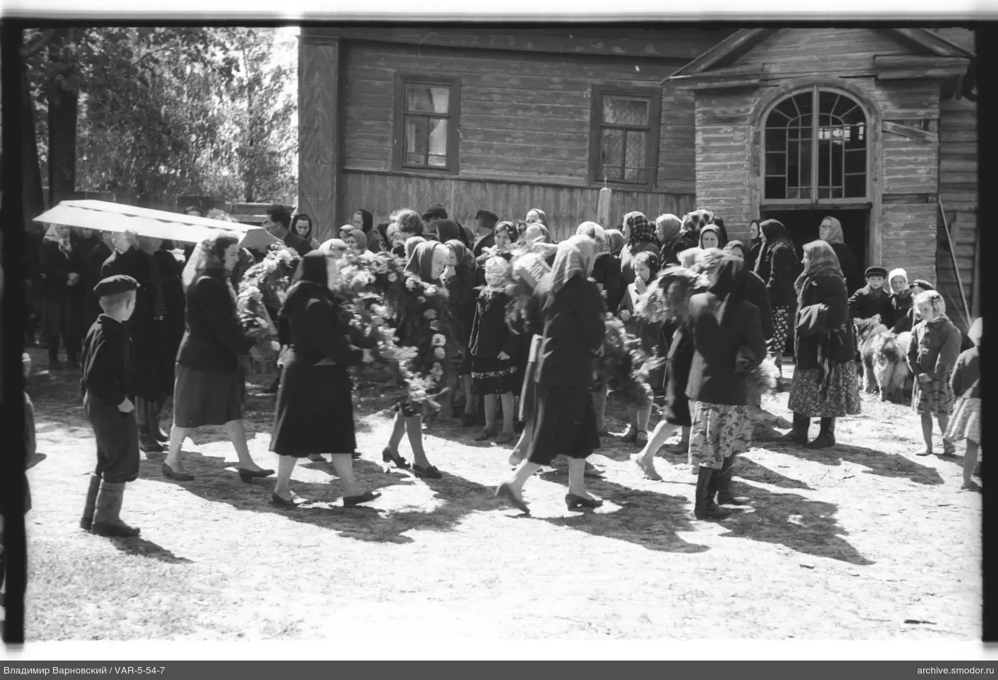 Gruppe von Frauen und Kindern mit Kränzen vor Holzgebäude